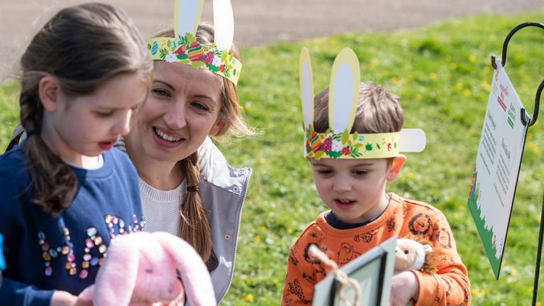 Two young children with woman, wearing bunny ears at an Easter activity point with grass in background